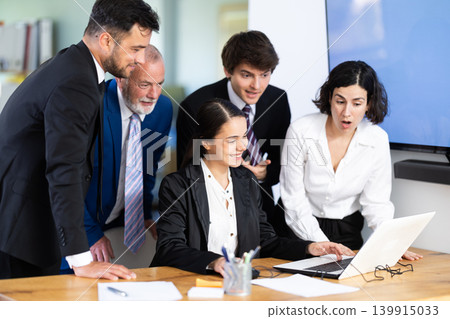 Cheerful team of co-workers working together, everyone looking at vacation photos on laptop by young girl and laughing in corporate office Cheerful team of co-workers working together, everyone looking at vacation photos on laptop by young girl and laughing in corporate office 139915033