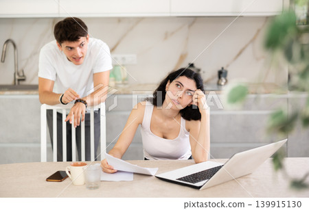 Sad girl works with laptop and paper documents and listens to reproaches of young guy Sad girl works with laptop and paper documents and listens to reproaches of young guy 139915130