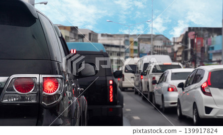 Tight rear view of black vehicle stopped on the asphalt road. City traffic during the day. Long queue of diverse many cars stretches ahead down a congested urban lane. Traffic congestion in Thailand. Tight rear view of black vehicle stopped on the asphalt road. City traffic during the day. Long queue of diverse many cars stretches ahead down a congested urban lane. Traffic congestion in Thailand. 139917824