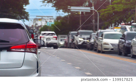 Vehicles are lined up in a heavy daytime traffic jam on a wide city road. A silver car is prominent in the foreground lane, with lush green trees lining the roadside in the background. 139917825