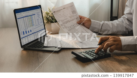 Accountant reviewing financial report and tax documents on laptop while using calculator for account analysis, real estate investment planning, expense management, and reporting workflow. Accountant reviewing financial report and tax documents on laptop while using calculator for account analysis, real estate investment planning, expense management, and reporting workflow. 139917847