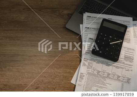 Calculator and pen placed on tax forms beside laptop on wooden desk for checking tax documents, accounting review, financial calculation, expense reporting, and tax filing workflow. Calculator and pen placed on tax forms beside laptop on wooden desk for checking tax documents, accounting review, financial calculation, expense reporting, and tax filing workflow. 139917859