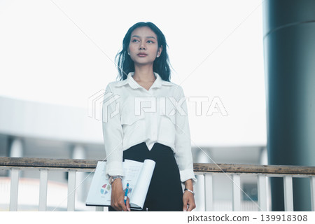 Low-angle portrait of a confident professional woman holding rolled documents and a clipboard in a modern office, expressing leadership, business planning, corporate readiness, and career success. Low-angle portrait of a confident professional woman holding rolled documents and a clipboard in a modern office, expressing leadership, business planning, corporate readiness, and career success. 139918308
