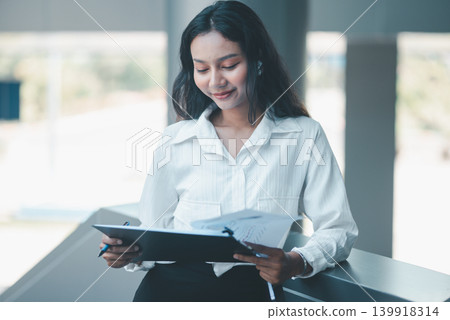 Professional woman reviewing business documents on a clipboard while leaning in a modern office, expressing focused analysis, report checking, organized workflow, and everyday corporate productivity. Professional woman reviewing business documents on a clipboard while leaning in a modern office, expressing focused analysis, report checking, organized workflow, and everyday corporate productivity. 139918314