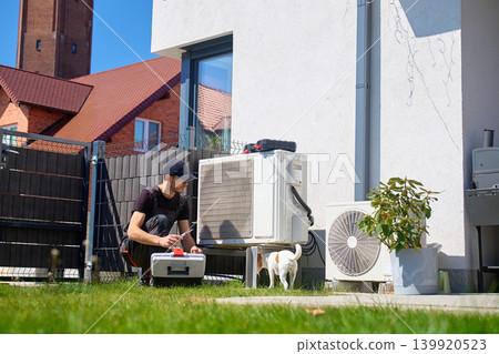 Man servicing outdoor heat pump with small dog Man servicing outdoor heat pump with small dog 139920523
