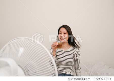 一位面帶微笑的女子在屋內一邊喝著大麥茶,一邊享受著風扇吹來的微風。 一位面帶微笑的女子在屋內一邊喝著大麥茶,一邊享受著風扇吹來的微風。 139921045