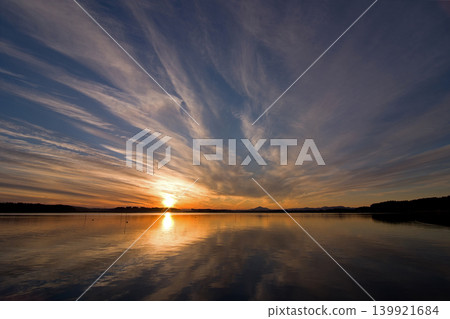 The sunset over Lake Hinuma in Ibaraki Prefecture. The orange glow of the sun, the white clouds, and the silhouette of Mount Tsukuba are reflected on the calm surface of the water, creating a beautiful scene. The sunset over Lake Hinuma in Ibaraki Prefecture. The orange glow of the sun, the white clouds, and the silhouette of Mount Tsukuba are reflected on the calm surface of the water, creating a beautiful scene. 139921684