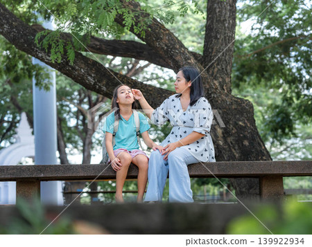 Happy Asian mother and her young daughter sitting under a big tree after school at the playground. Happy Asian mother and her young daughter sitting under a big tree after school at the playground. 139922934