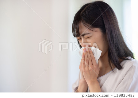A young Japanese woman blows her nose in a brightly lit living room. 139923482