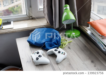 Two gaming controllers sit on a wooden desk next to a green lamp and stacks of books. 139923756