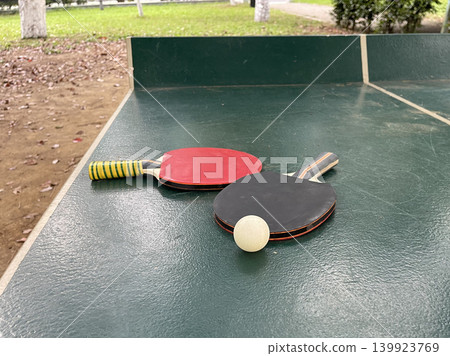 Two table tennis paddles and a ball are placed on a green table in a park during the day  139923769