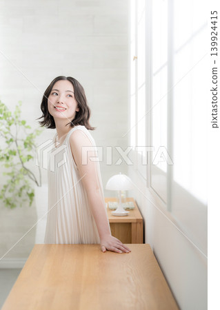 A vertical image of a smiling woman gazing out the window in a room bathed in natural light, representing beauty and a natural lifestyle. 139924415