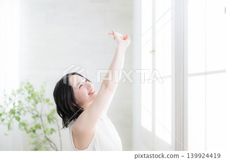An image of a healthy lifestyle of a woman waking up by the window, basking in the morning light, and stretching. An image of a healthy lifestyle of a woman waking up by the window, basking in the morning light, and stretching. 139924419