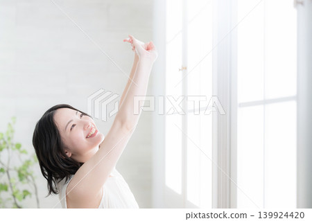 A healthy lifestyle image of a woman stretching while basking in the morning light by the window. A healthy lifestyle image of a woman stretching while basking in the morning light by the window. 139924420