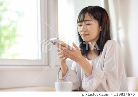 A young Japanese woman is using her smartphone at a table by the window. A young Japanese woman is using her smartphone at a table by the window. 139924670