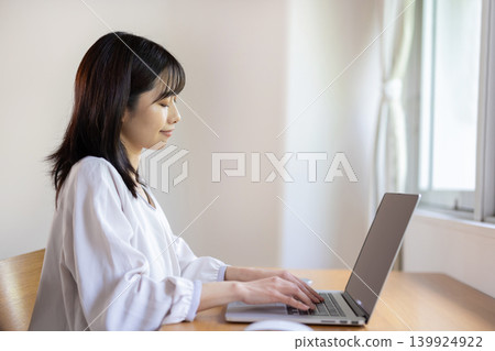 一位年輕的日本女性在明亮的窗邊桌子旁使用筆記型電腦工作。 139924922