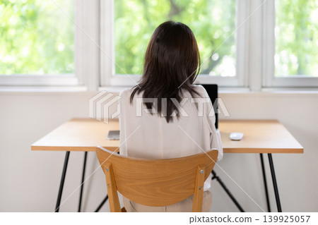 The back view of a woman working on her laptop at a table by the window. The back view of a woman working on her laptop at a table by the window. 139925057