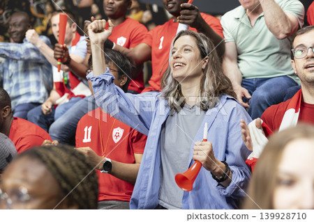 Woman football supporter cheering with red horn in red team stadium crowd during energetic live soccer match. Modern football culture with inclusion and equality, football is for everyone. 139928710