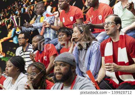 Red team football supporters watching tense live soccer match from stadium stands, mixed emotions, horns and anxious crowd reaction. Sports fans clenching fists speechless after missed goal. Red team football supporters watching tense live soccer match from stadium stands, mixed emotions, horns and anxious crowd reaction. Sports fans clenching fists speechless after missed goal. 139928712