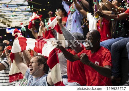 Excited football supporters celebrating with confetti in stadium stands, waving red scarves and cheering during live soccer match victory and team spirit. Ecstatic supporters celebrate champions team. Excited football supporters celebrating with confetti in stadium stands, waving red scarves and cheering during live soccer match victory and team spirit. Ecstatic supporters celebrate champions team. 139928717
