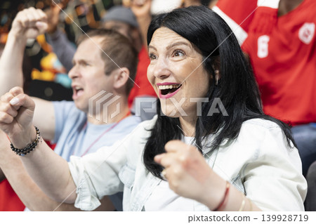 Ecstatic woman supporter celebrating big win during crucial football match after team scores the winning goal. Joyful fans create unforgettable stadium euphoria, jumping with raised fists. Ecstatic woman supporter celebrating big win during crucial football match after team scores the winning goal. Joyful fans create unforgettable stadium euphoria, jumping with raised fists. 139928719
