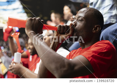 African american supporter with a vuvuzela leads synchronized fan chants in crowded stadium stands during live soccer match. Diverse sports fans celebrating and cheering, rhythmic noise. 139928720