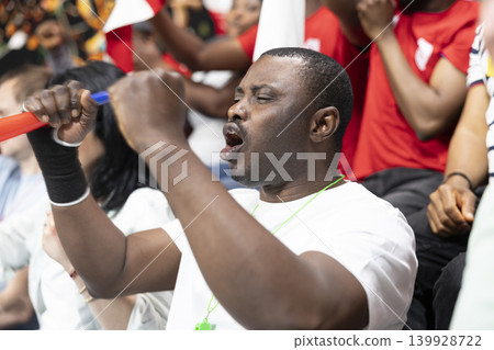 Happy black supporter celebrating football victory after the final whistle of a dramatic football match, jumping and screaming in the stadium stands with other fans. Ecstatic people raising fists. 139928722