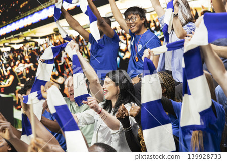 Singing blue team football fans waving scarves and cheering together in packed stadium stands during energetic live soccer match. Team spirit and celebration inside the packed stadium. Singing blue team football fans waving scarves and cheering together in packed stadium stands during energetic live soccer match. Team spirit and celebration inside the packed stadium. 139928733