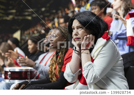 Mature female football supporter covering ears in loud stadium crowd, red scarf and anxious reaction during intense live soccer match. Inclusive fans with women in football, football community. Mature female football supporter covering ears in loud stadium crowd, red scarf and anxious reaction during intense live soccer match. Inclusive fans with women in football, football community. 139928734