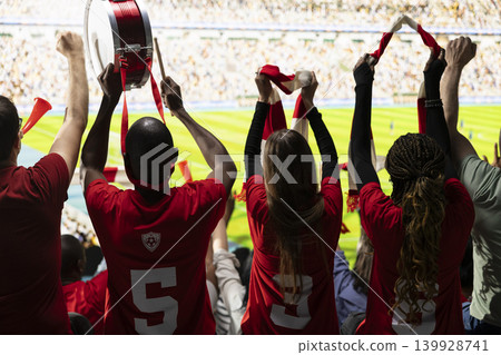 Back view of passionate supporters cheering in the stadium stands during football match filled with crowd noise and excitement. Diverse soccer ultras wave raised scarves, play fan drums. Back view of passionate supporters cheering in the stadium stands during football match filled with crowd noise and excitement. Diverse soccer ultras wave raised scarves, play fan drums. 139928741