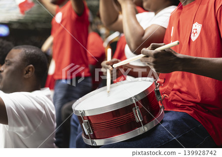 Energetic football supporter beating a percussion drum in the terrace stands, leading chanting fans during live soccer match. Hardcore supporters in the fan section celebrate with loud stadium noise. Energetic football supporter beating a percussion drum in the terrace stands, leading chanting fans during live soccer match. Hardcore supporters in the fan section celebrate with loud stadium noise. 139928742
