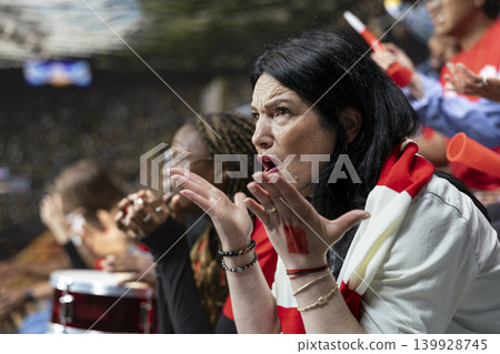 Emotional mature female football fan reacting with open hands in disbelief during crucial soccer match moment from stadium stands. Dejected supporters of the losing team remain stunned. Emotional mature female football fan reacting with open hands in disbelief during crucial soccer match moment from stadium stands. Dejected supporters of the losing team remain stunned. 139928745