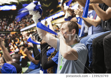 Passionate male football supporter blowing blue vuvuzela in crowded stadium stands during loud live soccer match atmosphere. Diverse football ultras create intense crowd noise, energy. Passionate male football supporter blowing blue vuvuzela in crowded stadium stands during loud live soccer match atmosphere. Diverse football ultras create intense crowd noise, energy. 139928749