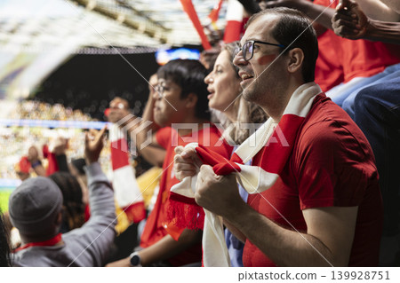 Anxious football supporters watching a decisive final attack during match from packed stadium stands with red scarves and fan tension. Supporter holding breath with nervous anticipation. Anxious football supporters watching a decisive final attack during match from packed stadium stands with red scarves and fan tension. Supporter holding breath with nervous anticipation. 139928751