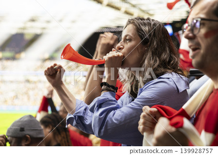 Passionate female football fan blowing a red vuvuzela while cheering in crowded stadium stands during an intense live soccer match. Diverse multigenerational supporters in the fan section. Passionate female football fan blowing a red vuvuzela while cheering in crowded stadium stands during an intense live soccer match. Diverse multigenerational supporters in the fan section. 139928757