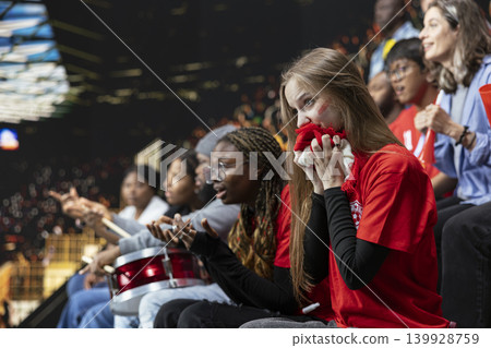 Anxious young football supporter biting red scarf in crowded stadium stands during tense live game, modern football with women empowerment and inclusion. Suspense and anticipation. 139928759