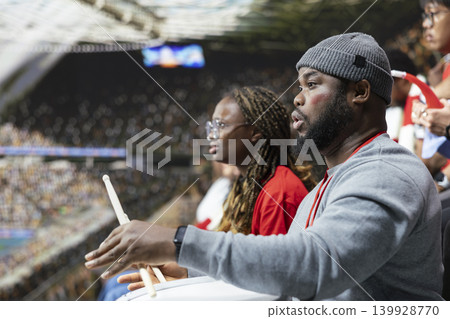 Focused football supporters watching tense live soccer match from stadium stands, side view with fan drum and anxious crowd atmosphere. Passionate fans holding breath under match suspense. Focused football supporters watching tense live soccer match from stadium stands, side view with fan drum and anxious crowd atmosphere. Passionate fans holding breath under match suspense. 139928770