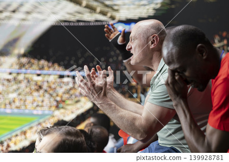 Frustrated male football fan pleading with open hands while another supporter covers face during tense stadium match reaction. Silent crowd reacting to last minute loss after final whistle. Frustrated male football fan pleading with open hands while another supporter covers face during tense stadium match reaction. Silent crowd reacting to last minute loss after final whistle. 139928781