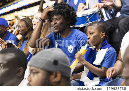 Excited young boy with vuvuzela watching live soccer match with parent in packed stadium, surrounded by crowd energy and passionate supporters. Black family cheering for national team, bonding. Excited young boy with vuvuzela watching live soccer match with parent in packed stadium, surrounded by crowd energy and passionate supporters. Black family cheering for national team, bonding. 139928786