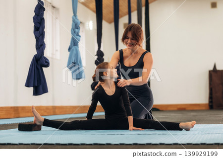 Busy by doing exercises. Female instructor helping a child practice yoga in a fitness studio Busy by doing exercises. Female instructor helping a child practice yoga in a fitness studio 139929199