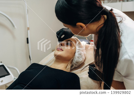 In black gloves. Female cosmetologist performing a facial skincare procedure using a cosmetic device in a beauty clinic In black gloves. Female cosmetologist performing a facial skincare procedure using a cosmetic device in a beauty clinic 139929802