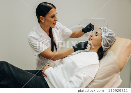 Client is lying down. Female cosmetologist performing a facial skincare procedure using a cosmetic device in a beauty clinic Client is lying down. Female cosmetologist performing a facial skincare procedure using a cosmetic device in a beauty clinic 139929811