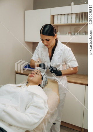 In black gloves. Female cosmetologist performing a facial skincare procedure using a cosmetic device in a beauty clinic In black gloves. Female cosmetologist performing a facial skincare procedure using a cosmetic device in a beauty clinic 139929815