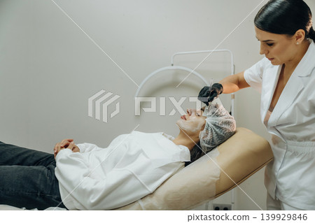 Dermatology conception. Female cosmetologist performing a facial skincare procedure using a cosmetic device in a beauty clinic Dermatology conception. Female cosmetologist performing a facial skincare procedure using a cosmetic device in a beauty clinic 139929846