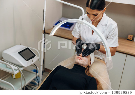 White cream, quality product. Female cosmetologist performing a facial skincare procedure using a cosmetic device in a beauty clinic White cream, quality product. Female cosmetologist performing a facial skincare procedure using a cosmetic device in a beauty clinic 139929889