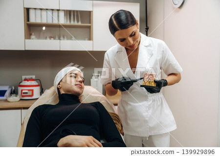 Talking, showing what's under the skin. Female cosmetologist performing a facial skincare procedure using a cosmetic device in a beauty clinic Talking, showing what's under the skin. Female cosmetologist performing a facial skincare procedure using a cosmetic device in a beauty clinic 139929896