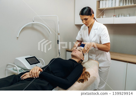 Female cosmetologist performing a facial skincare procedure using a cosmetic device in a beauty clinic Female cosmetologist performing a facial skincare procedure using a cosmetic device in a beauty clinic 139929914