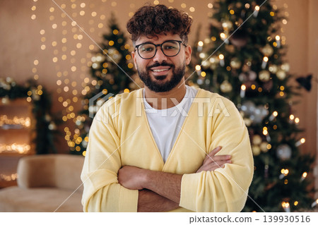 Arms crossed, standing and smiling. Man posing in front of a Christmas tree in a festive home setting Arms crossed, standing and smiling. Man posing in front of a Christmas tree in a festive home setting 139930516