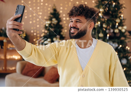 Making a selfie by using smartphone. Man posing in front of a Christmas tree in a festive home setting Making a selfie by using smartphone. Man posing in front of a Christmas tree in a festive home setting 139930525