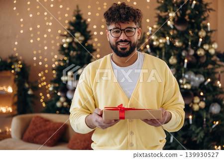 Gift box is in hands. Man posing in front of a Christmas tree in a festive home setting Gift box is in hands. Man posing in front of a Christmas tree in a festive home setting 139930527